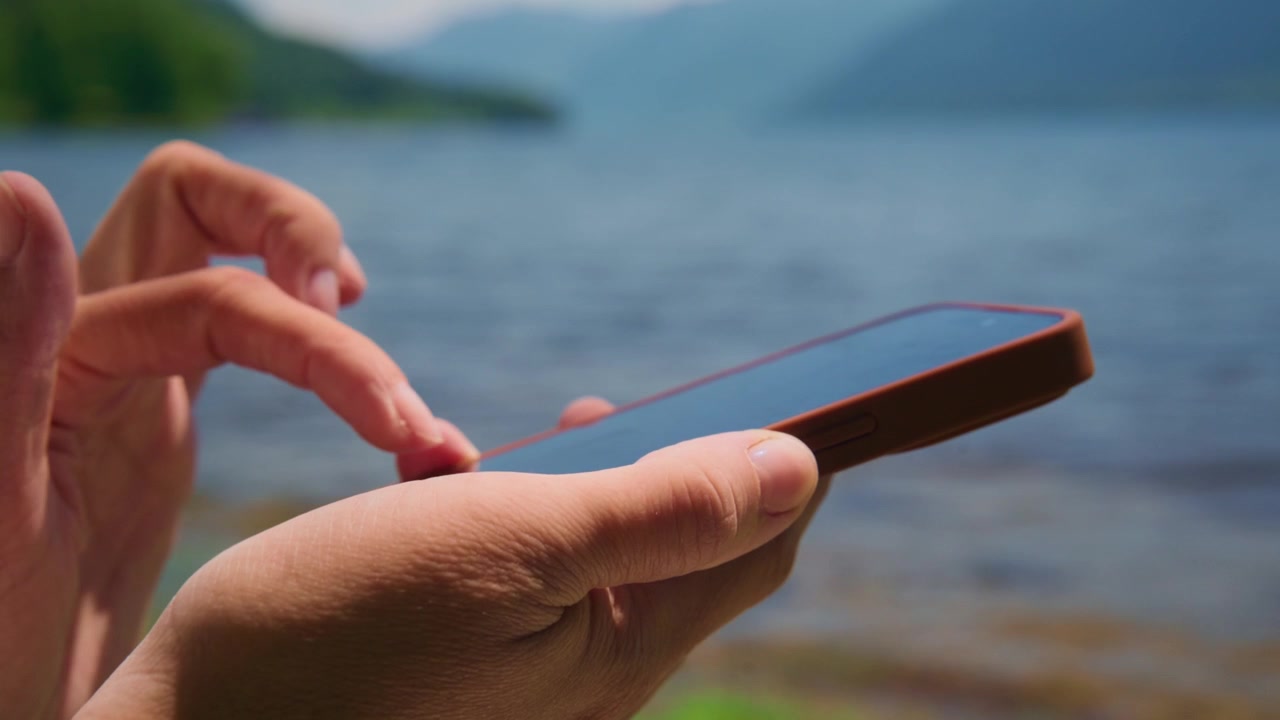 Mani che usano il telefono vicino a un lago. Lo sfondo mostra acqua calma e colline verdi distanti. La persona appare rilassata e concentrata sul dispositivo. Ideale per temi di viaggio, tecnologia o comunicazione all'aperto.