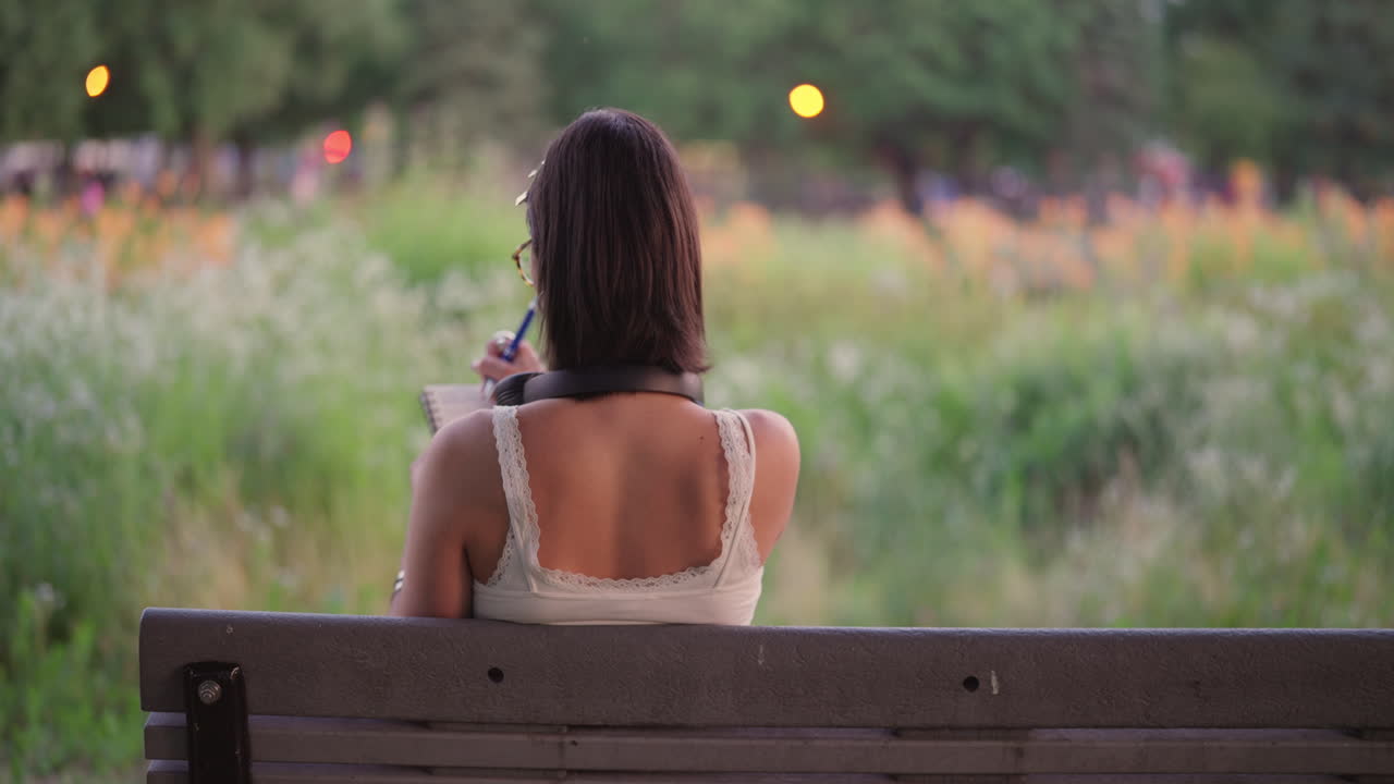 Back view of woman on bench with headphones around neck journaling or drawing in front of field