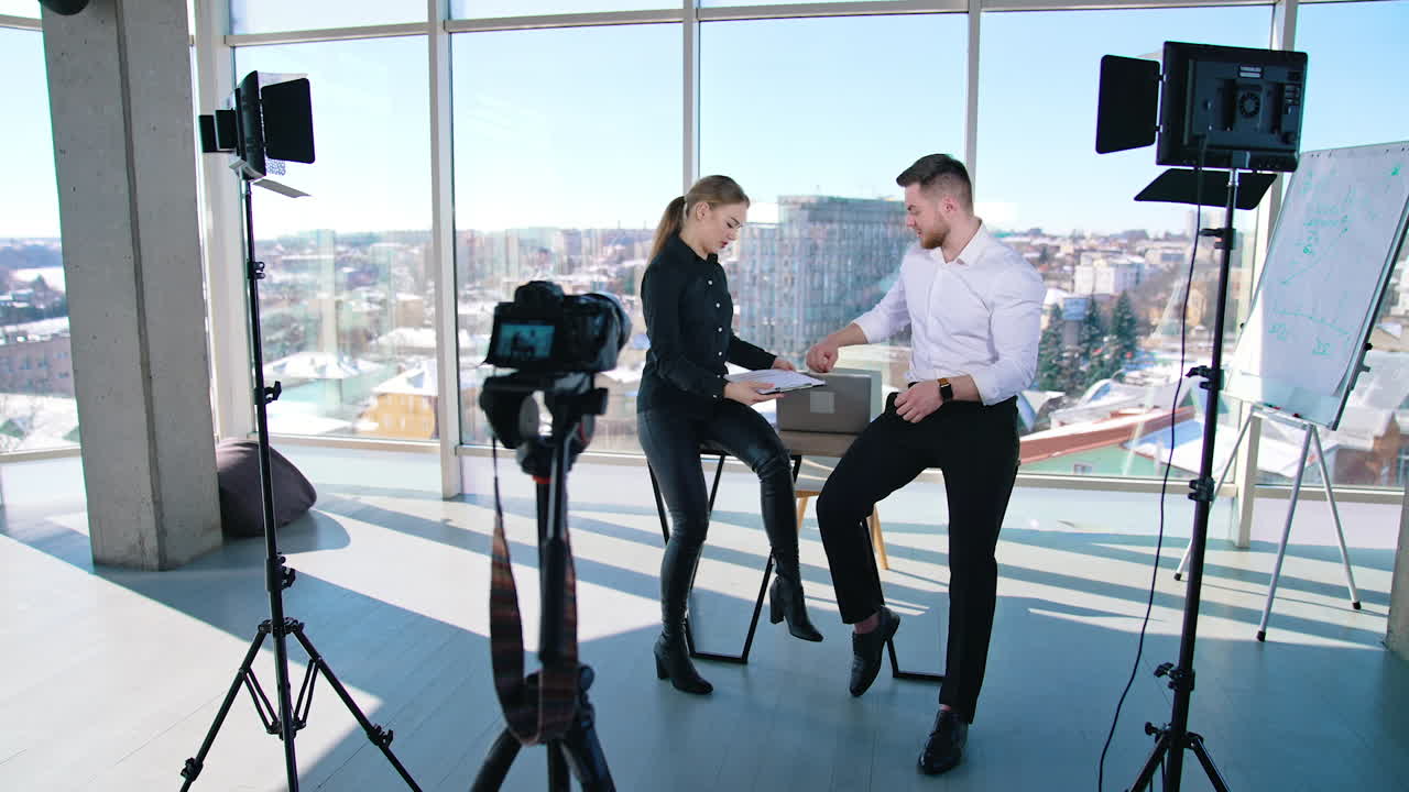 Young entrepreneurs in front of professional camera. Pretty woman and handsome guy sitting in office room and speaking about their new startup on camera.