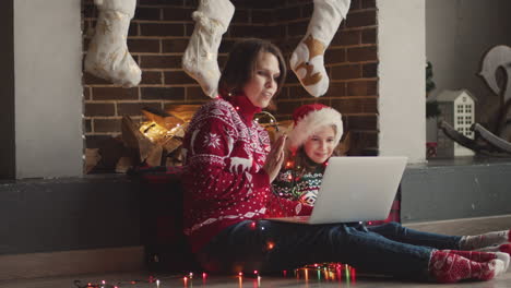 feliz madre e hija en suéteres navideños y con luces alrededor del cuello sentadas en el suelo mientras usan una computadora portátil