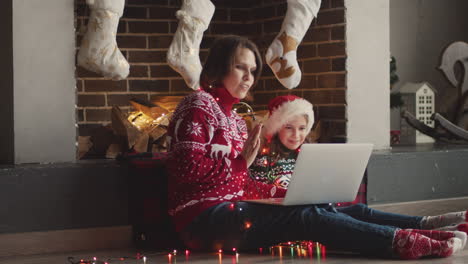 happy mother and daughter in christmas sweaters and with lights around neck sitting on the floor while using laptop computer