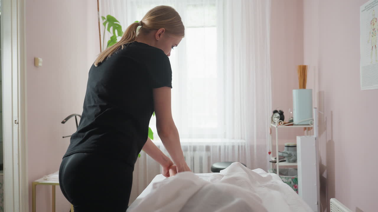 Massage therapist in black attire pressing client under foot using fingers in well-lit room with soft curtains, indoor plant, and gentle decoration creating calm spa environment for therapy