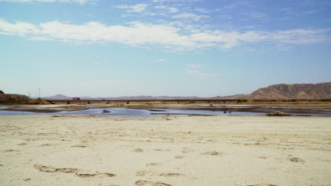 On-a-sunny-day,-the-yellow-sand-of-Zorritos-beach-in-Tumbes,-in-Peru-can-be-seen-with-multiple-cars-driving-in-the-distance