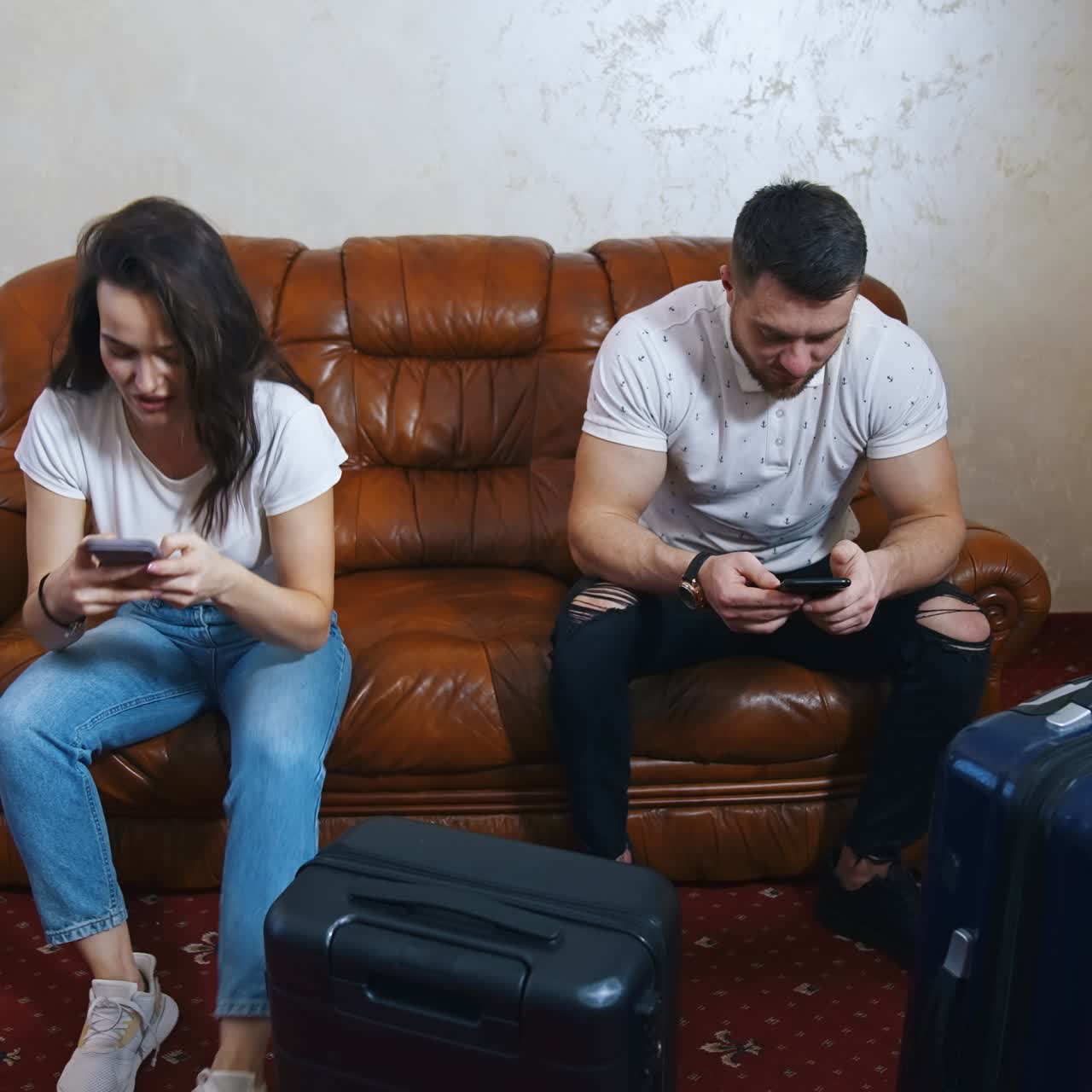 Tourist couple on sofa with travel bags nearby. Young woman and man using phones while waiting for transport at home. Packed suitcases in the hotel