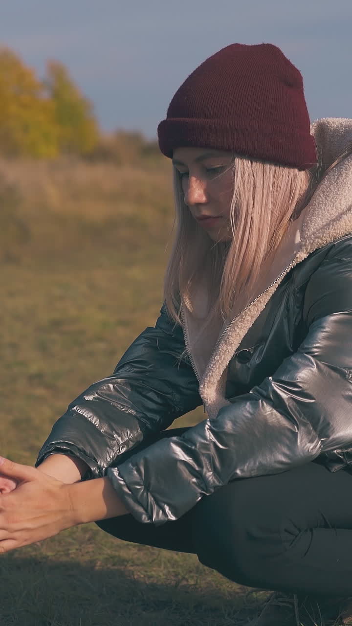 blonde girl tourist in warm jacket heats hands at burning bonfire at camp against yellow trees in autumn morning