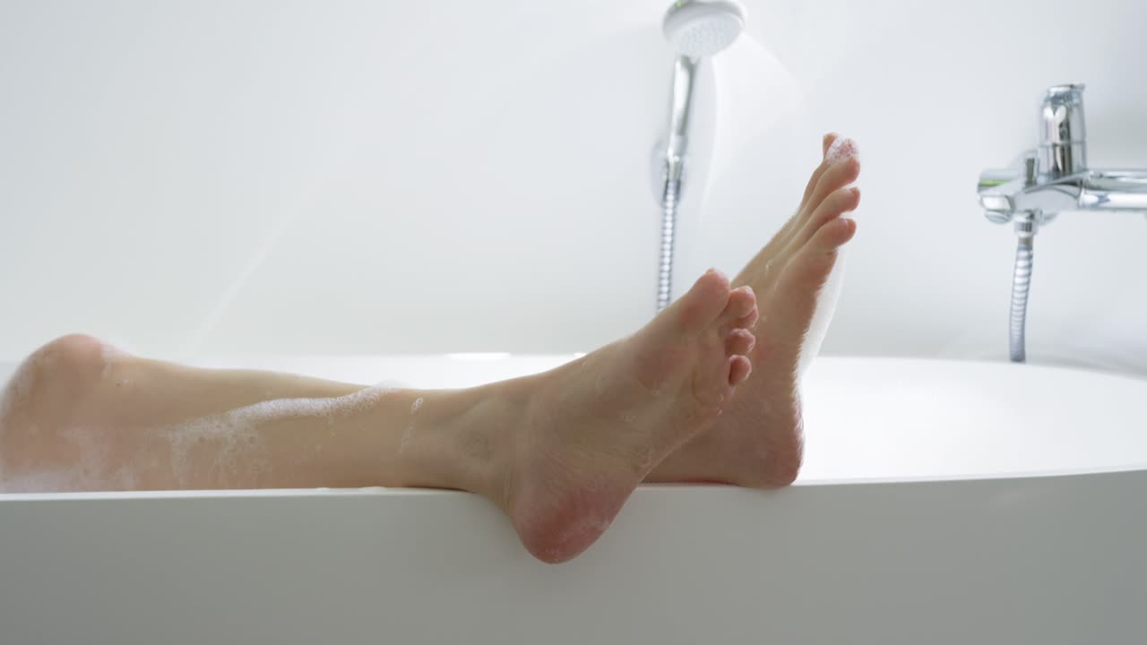 Piernas y pies de mujer flexionándose mientras se enjuaga en el borde de la bañera, mostrando gotas del cabezal de la ducha