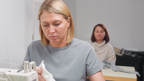 two female clothing designers sit in bright workspace, one focusing on sewing machine while other glances around casually, both engaged in discussion beside their fabric and equipment