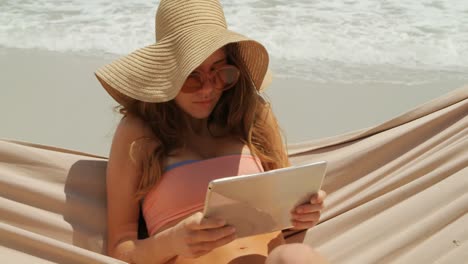 vista frontal de una mujer caucásica usando una tableta digital en una hamaca en la playa 4k
