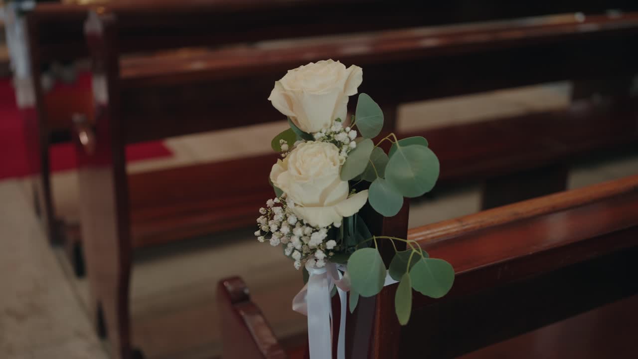 Close up of white roses, eucalyptus leaves, and ribbon tied to a wooden church pew for a wedding ceremony