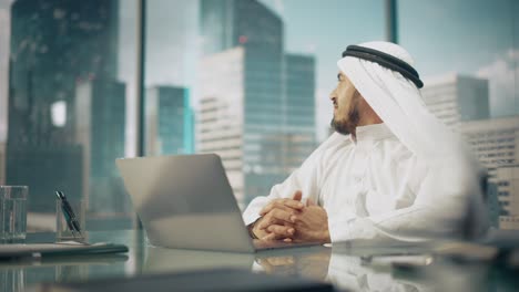 successful arab businessman in white traditional outfit sitting in office and working on laptop computer. business manager planning corporate strategy. saudi, emirati, arab businessman concept.