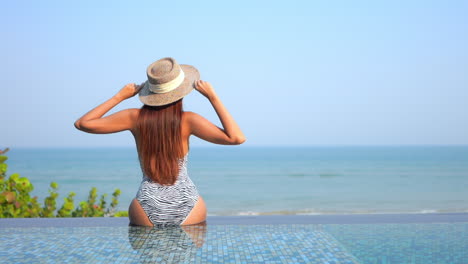 A-woman-with-her-back-to-the-camera-is-sitting-on-the-edge-of-an-infinity-pool-while-facing-the-ocean-raises-her-arms-in-joy