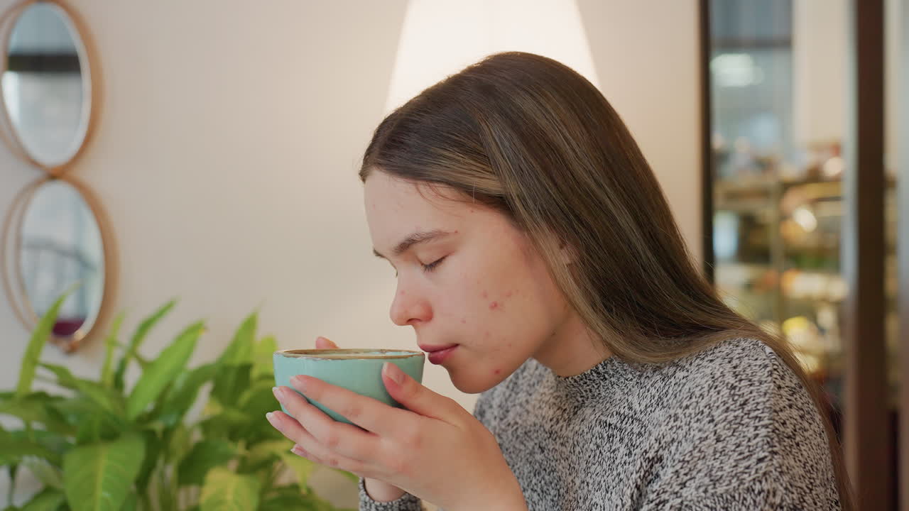 Young lady gently smelling aroma from coffee held with both hands satisfied expression indoor cozy setting soft lamp light potted plant mirror reflecting blurred people in background