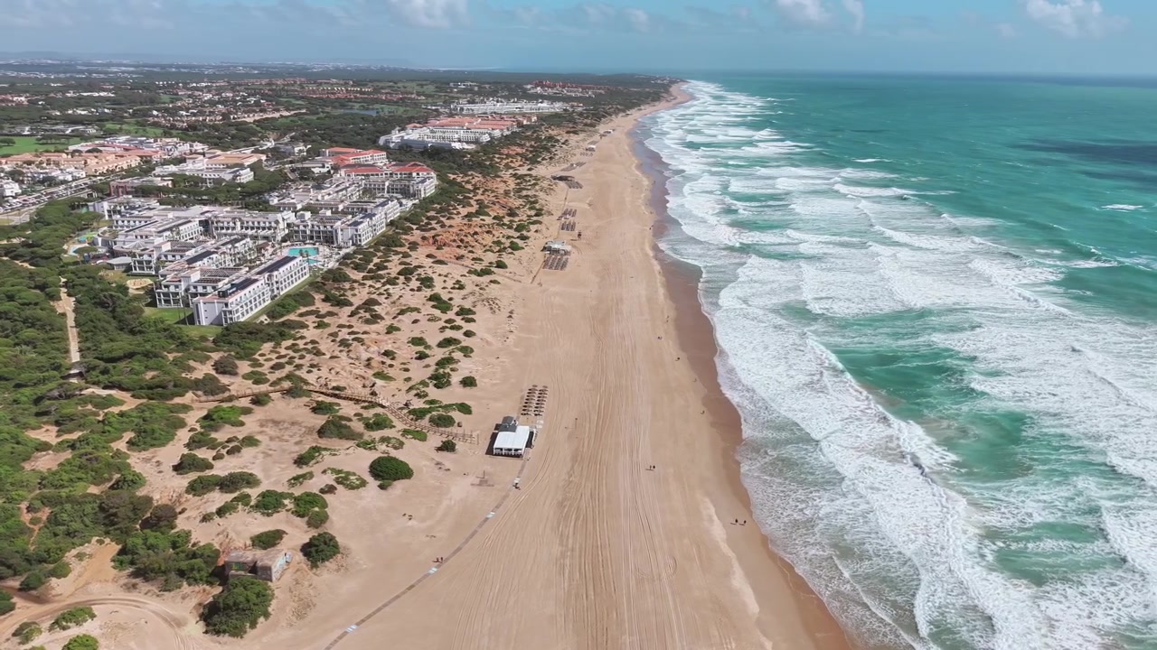 Órbita de dron cerca de hoteles y complejos turísticos que bordean una larga playa de arena, Playa de la Barrosa, en la costa atlántica cerca de Cádiz, mientras las olas rompen bajo cielos diurnos despejados, Cádiz, Andalucía, España