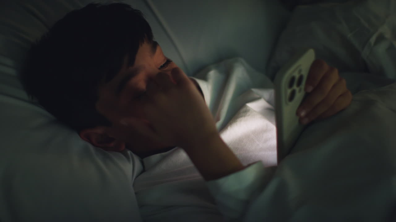 집에서 침실에서 피곤한 어린 소년은 밤에 문자 메시지를 보내기 위해 휴대 전화를 사용하여 침대에 누워 있습니다.