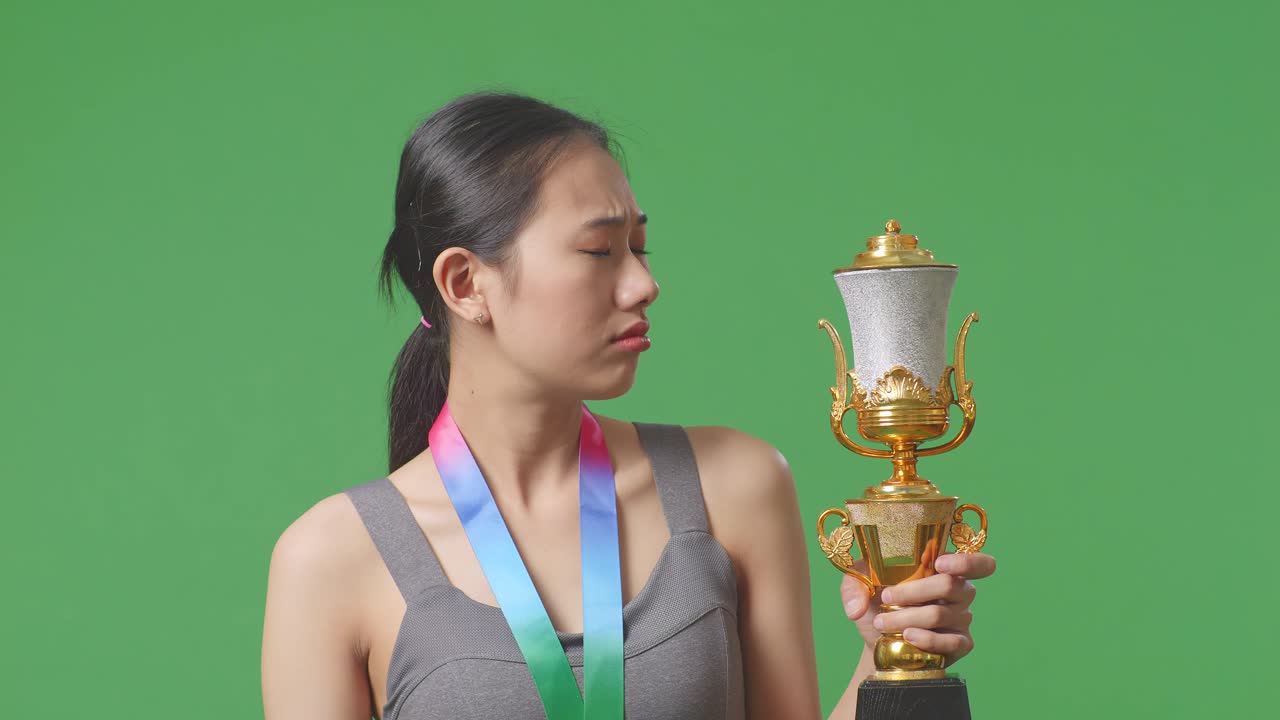 금메달을  아시아 여성이 트로피를 쳐다보며 머리를 흔들며 불만을 표하며 스튜디오의 녹색 스크린 배경에서 첫 번째 우승자로 승리합니다.