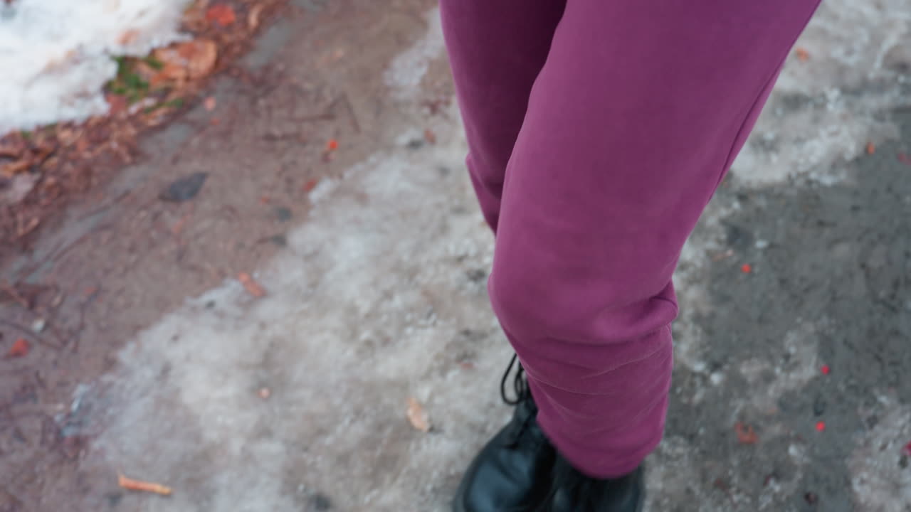 primer plano de la parte inferior del cuerpo con pantalones marrones y botas negras caminando por un camino de invierno áspero cubierto de follaje esparcido y manchas heladas, nieve y mezcla de tierra en el suelo desigual