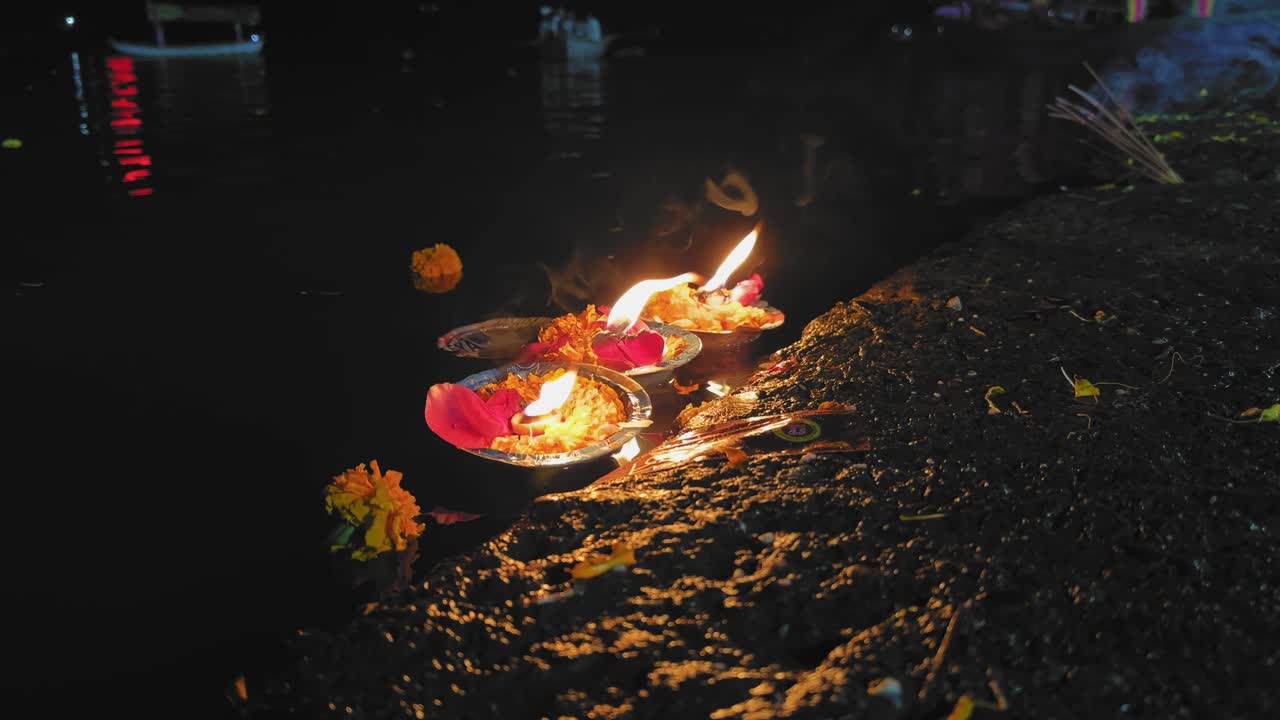 Diyas acesos colocados numa margem de rio molhada durante um ritual indiano brilham suavemente à noite, rodeados por pétalas de calêndula enquanto reflexos cintilantes se movem pela água sagrada em devoção.