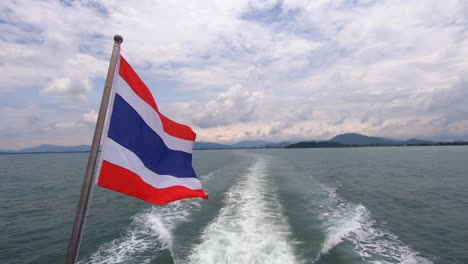 thai flag waving in wind on a boat ride with ocean wake pushing through the sea and islands in the background