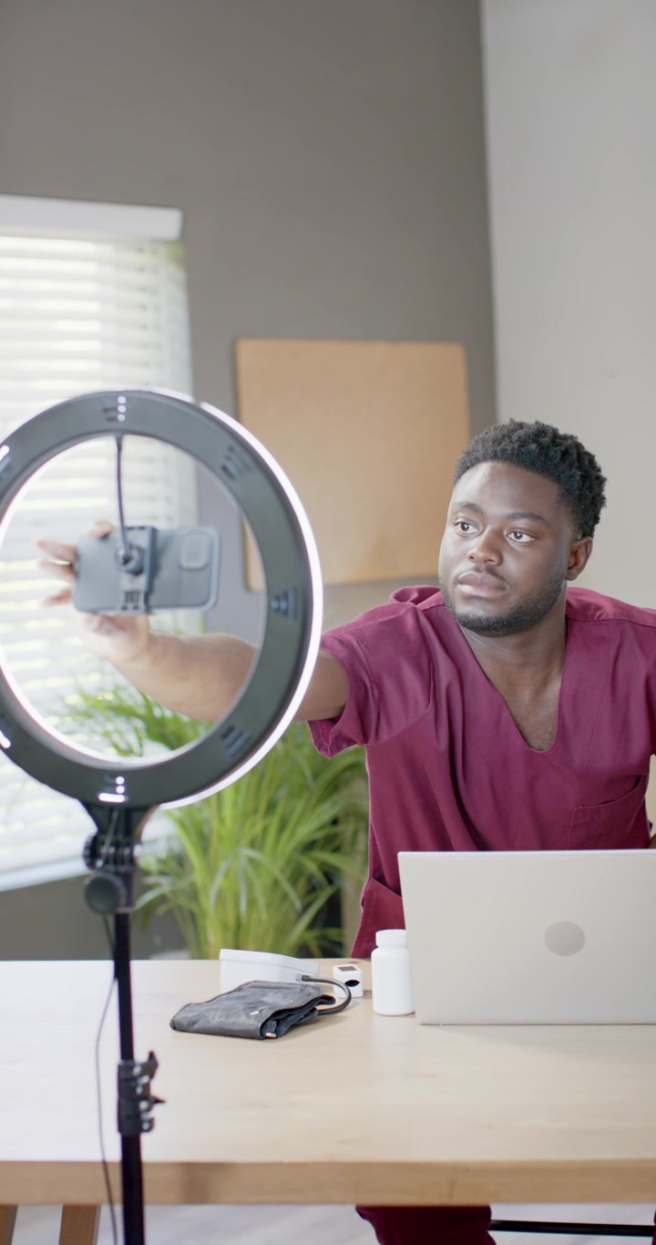 Video verticale: Uomo africano in camice medico che sistema un telefono inclinato in una ring light sulla scrivania per la registrazione