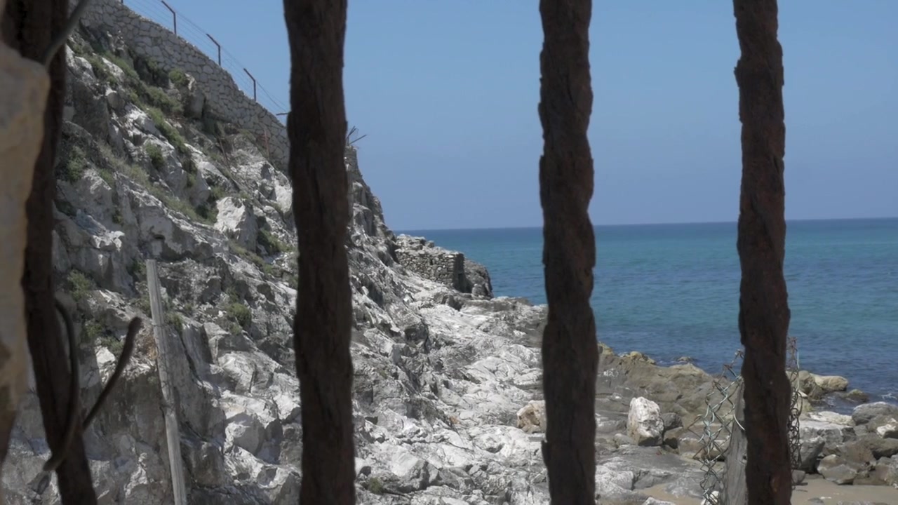 Havet avsløres gjennom rustne gitter. Vakkert landskap med blått hav og klipper.