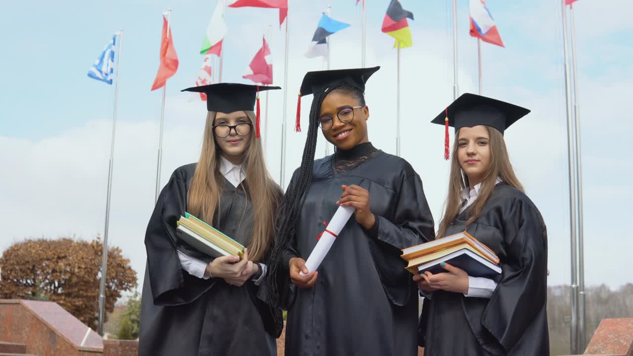 tres jóvenes graduadas universitarias de diferentes nacionalidades sonríen y se paran juntas sosteniendo libros y un diploma en sus manos contra muchas banderas de diferentes países