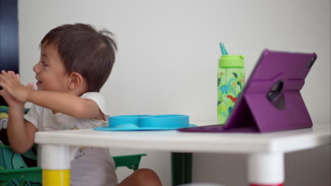 Lindo-Bebé-Latino-Aplaudiendo-Y-Disfrutando-De-Una-Película-En-Una-Tableta-Morada-Sentada-En-Una-Mesa-Blanca