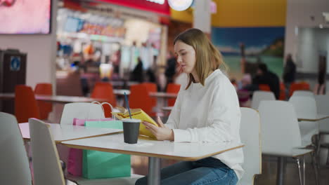 mujer joven sentada en un centro comercial ocupado casualmente leyendo un libro, rodeada por la vibrante atmósfera del centro comercial, bolsas de compras de colores en la mesa, y fondo borroso con personas y mostradores de comida rápida visibles