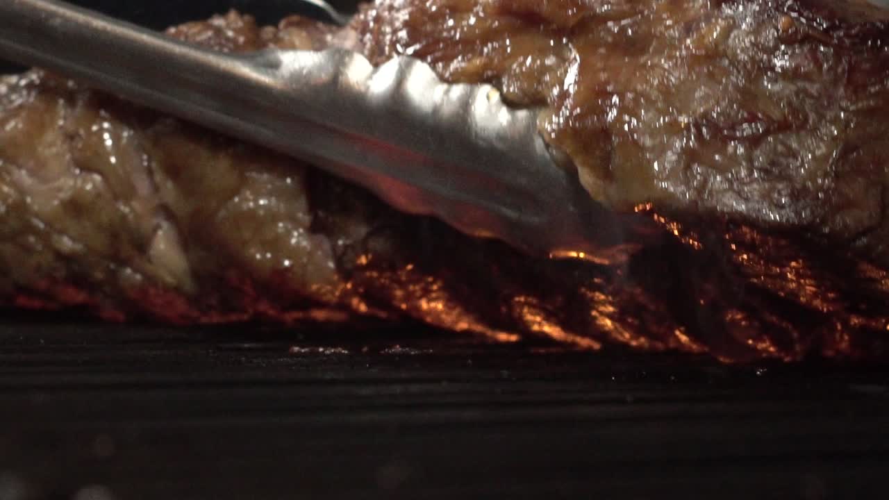 A tasty tomahawk steak on the cooking grill fire, extreme close up slow motion shot. Using a tong to turning the piece of meat over the grilling flame.