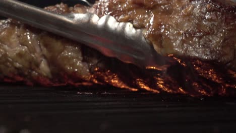 a tasty tomahawk steak on the cooking grill fire, extreme close up slow motion shot. using a tong to turning the piece of meat over the grilling flame.