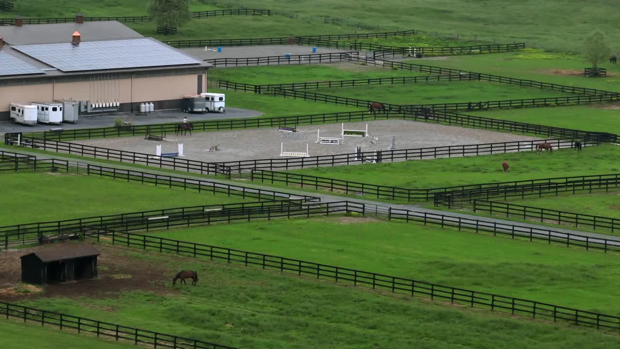 Equestrian on horse at american horse farm with grass fields. Aerial wide shot. Modern solar panels on horse barn.