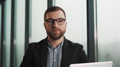 Retrato-De-Un-Joven-Sonriente-Con-Gafas-En-El-Fondo-De-Una-Oficina-Con-Ventanas-Panorámicas