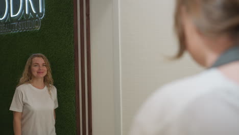 smiling customer with long blond hair wearing white shirt enters beauty studio and walks past green textured wall toward reception, warmly greeted by staff in calm and modern interior space