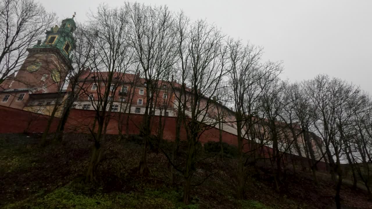 Visitatori camminano vicino a una collina con alberi che si affaccia su un sito storico. Il cielo è nuvoloso e la struttura mostra segni del tempo e della storia. La natura circonda l'area.