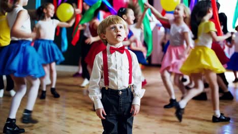 little boy wearing suspenders and bow tie watching other children dancing at a party, they're wearing colorful dresses and holding ribbons, he's standing still in the middle of the dance floor