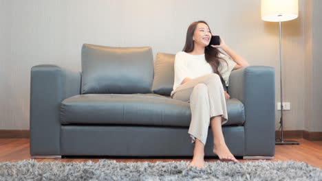 Sexy-Asiatische-Frau,-Die-Auf-Einem-Handy-Spricht,-Während-Sie-Zu-Hause-Auf-Dem-Sofa-Sitzt-Und-Lässige-Kleidung-Trägt,-Flirtender-Gesichtsausdruck