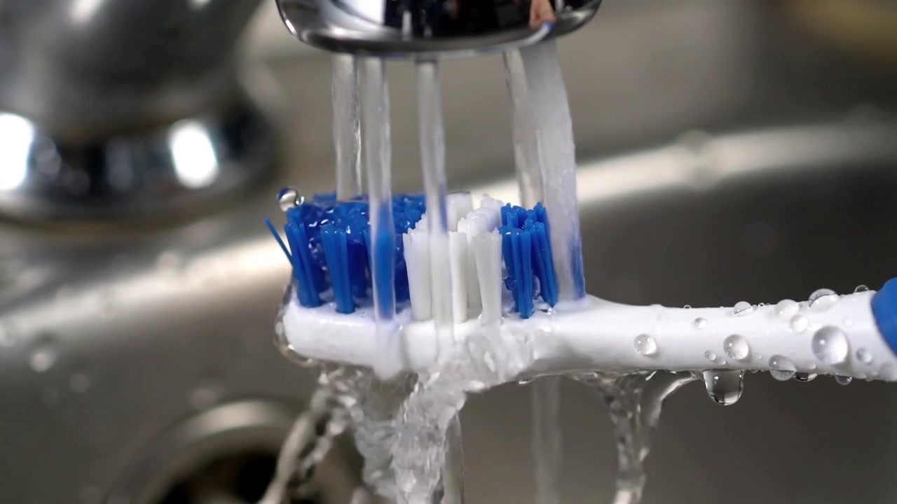 Faucet turning on, chrome streams rinsing white toothbrush at bathroom sink, removing blue paste