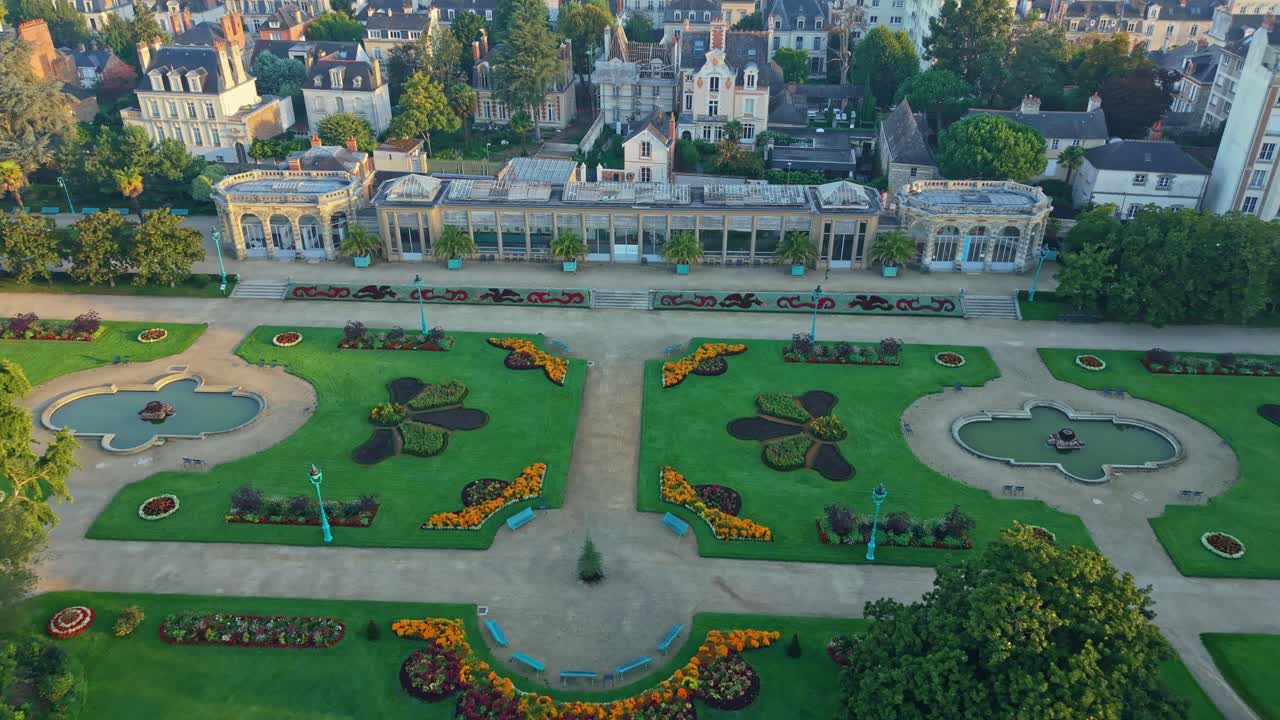 Drone shot descending toward Parc du Thabor in Rennes, revealing French-style gardens, paths, fountains, surrounding buildings and warm sunrise light