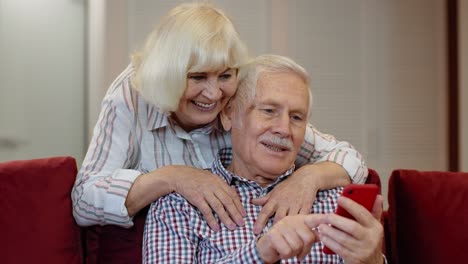 Glückliches-Lächelndes-älteres-Paar-Mit-Mobiltelefon-Zu-Hause.-Ausruhen-Auf-Dem-Sofa-Im-Gemütlichen-Wohnzimmer