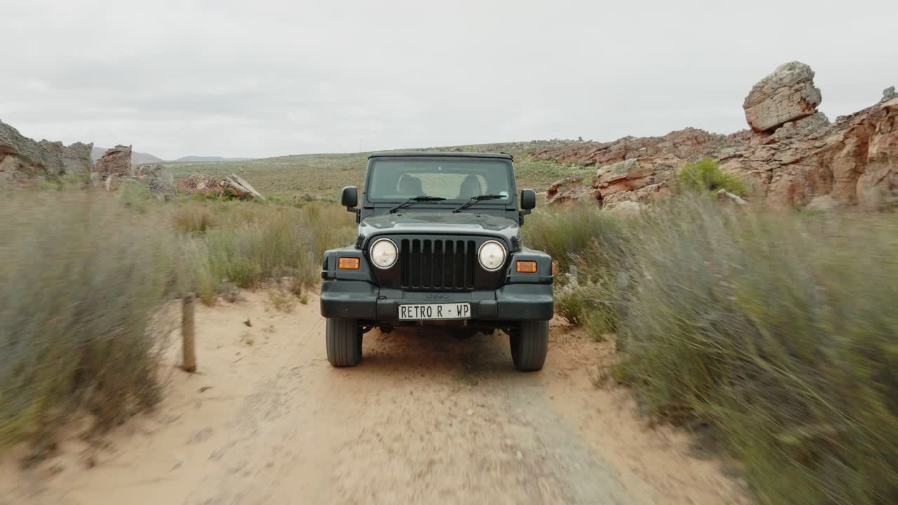 Drone flies fast backwards over ground in front of a black off-road vehicle over desert landscape and through rock formations in Cederberg Wilderness Area in South Africa