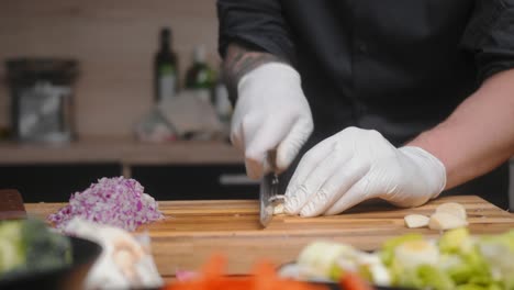 Fresh-garlic-being-cut-on-a-wooden-board-by-professional-chef-in-an-elegant-black-shirt-with-tattoos-and-white-gloves