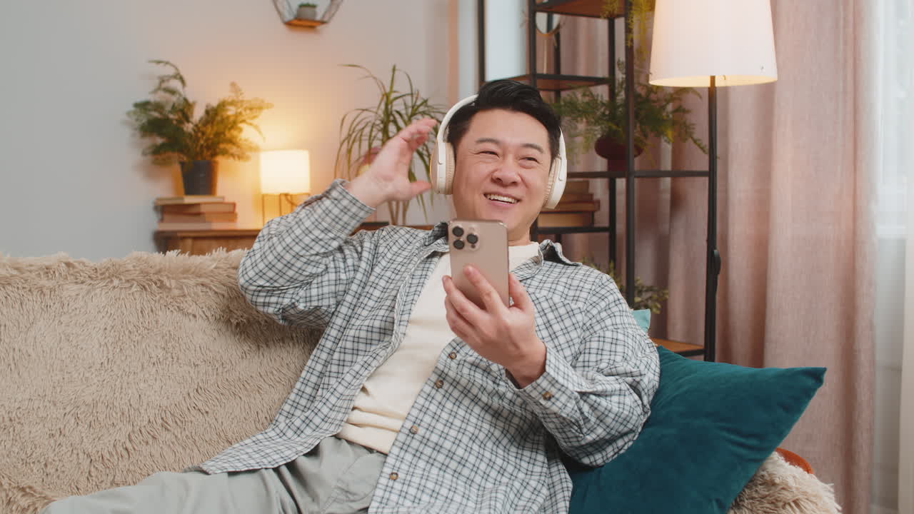 무선 헤드폰을 착용한 행복하고 즐거운 젊은 남자가 집에서 소파에 앉아 휴식을 취하고 있습니다.