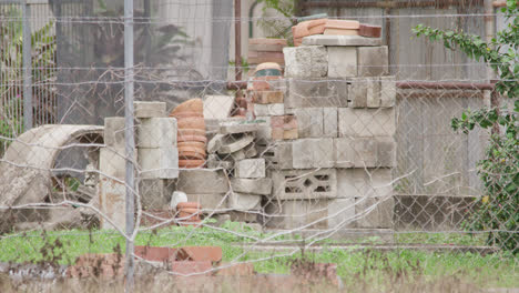 pan left, a pile of worksite rubble of concrete cinder blocks and terracotta bricks behind chain link wire fencing and barbed wire with dead vine entwined