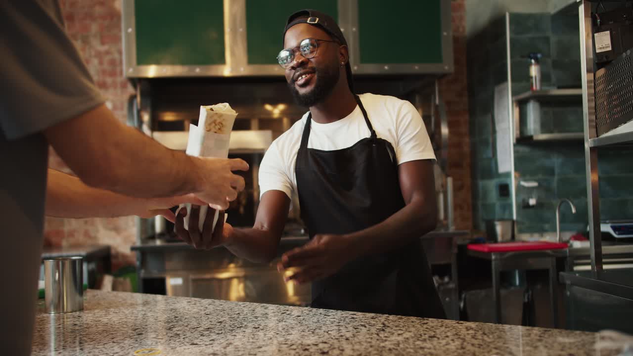 The Black person gives the order to the visitor. Fast food worker in glasses smiling and serving a visitor