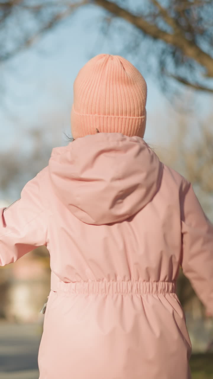padre en chaqueta marrón y vaqueros negros juega saltando con su hija en una gorra rosa y chaqueta, ambos disfrutando de un día soleado en el parque