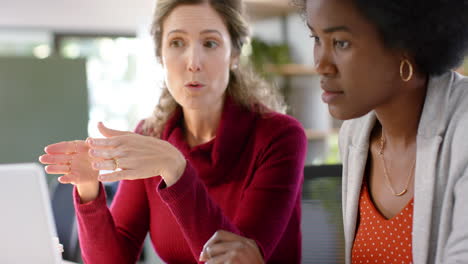 diverse female creative colleagues in discussion using laptop in office, slow motion