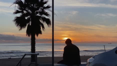 silueta de un hombre fumando contra una puesta de sol naranja, un joven fuma un cigarrillo con la playa y las olas detrás