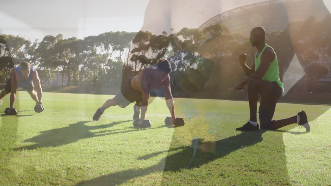 Exercising with dumbbells on grass, people near trees and stadium in background