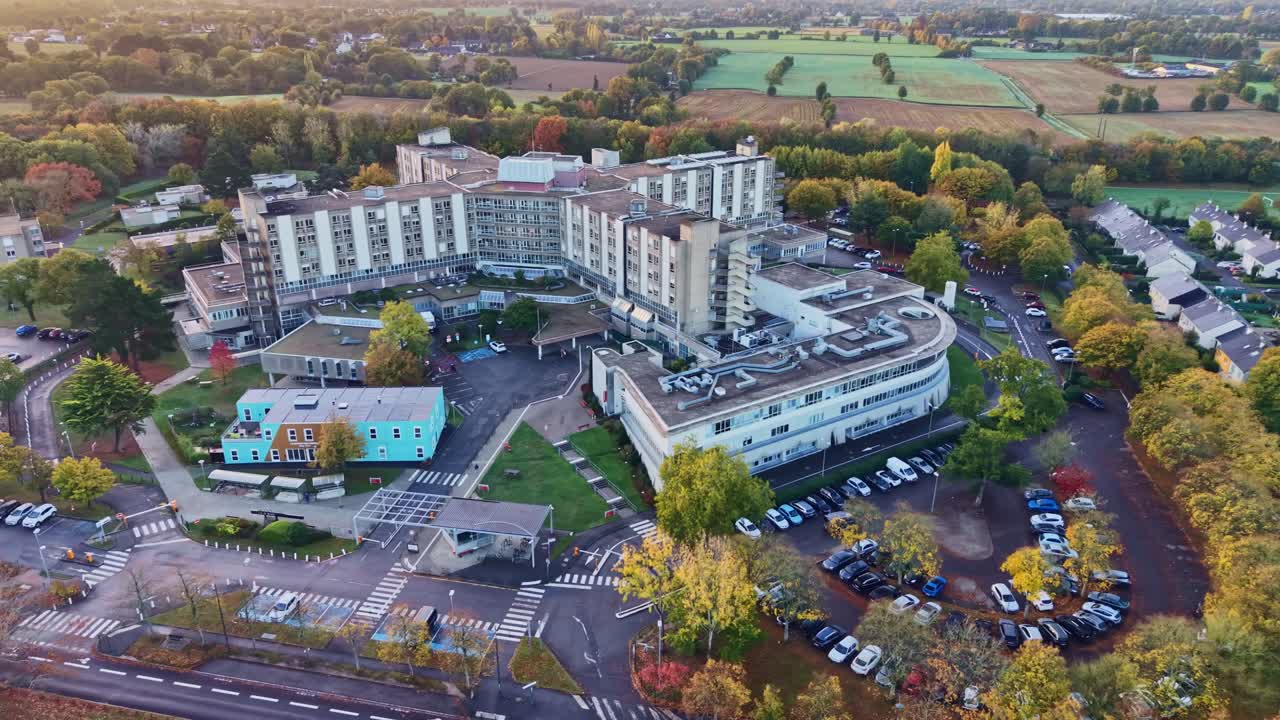 Drone establishing shot of CHU Hôpital Sud in Rennes, France, showing the sprawling hospital complex surrounded by tree-lined roads, parking areas, and green countryside on a sunny autumn morning