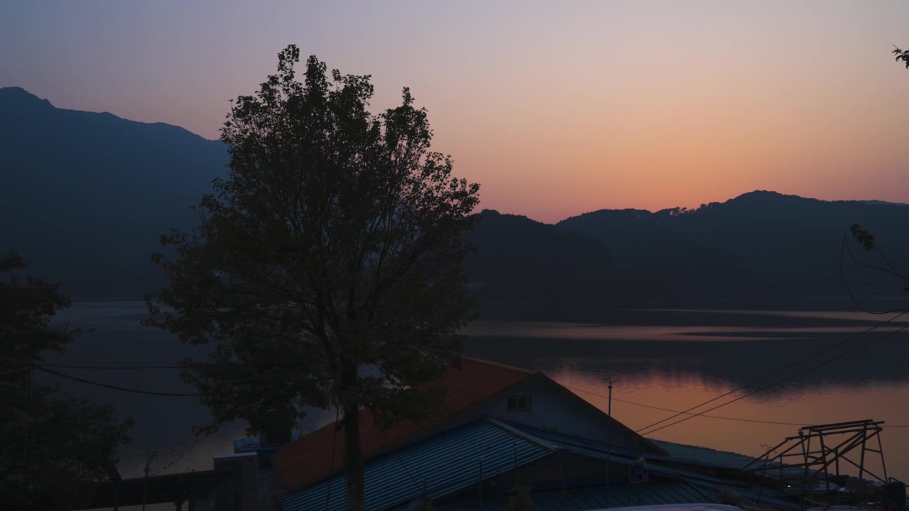 Auringonlasku hiipuu järvenrantapuun taakse vuoren juurella, kun tyyni vesi heijastaa lämpimiä iltasävyjä, Naejangsan, Etelä-Korea
