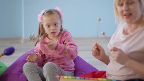 juguetes de aprendizaje, niña con síndrome de down con campanas jugando con una mujer con un xilófono durante la lección de música sentada en sillas de bolsa de frijol
